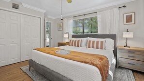 Un dormitorio con una cama, mesitas de noche, ventilador de techo, ventana con persianas y una puerta.