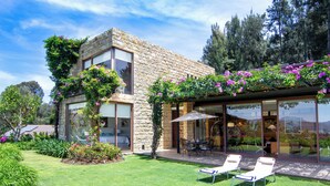 Una casa moderna con fachada de piedra, un invernadero de cristal y un césped cuidado con dos sillones de descanso.