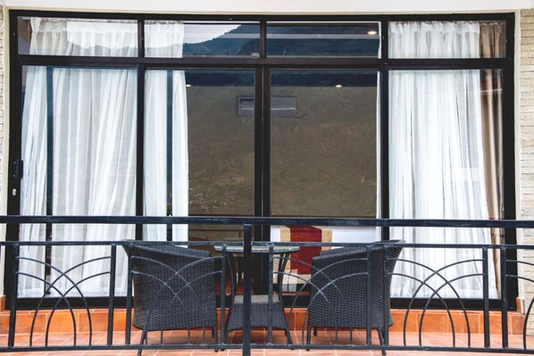 Un balcón con mesa y sillas, ventanas amplias con cortinas y vistas a un paisaje montañoso.