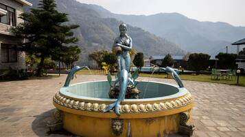 Una fuente con una estatua de sirena en una base circular, rodeada por una zona pavimentada con montañas al fondo.
