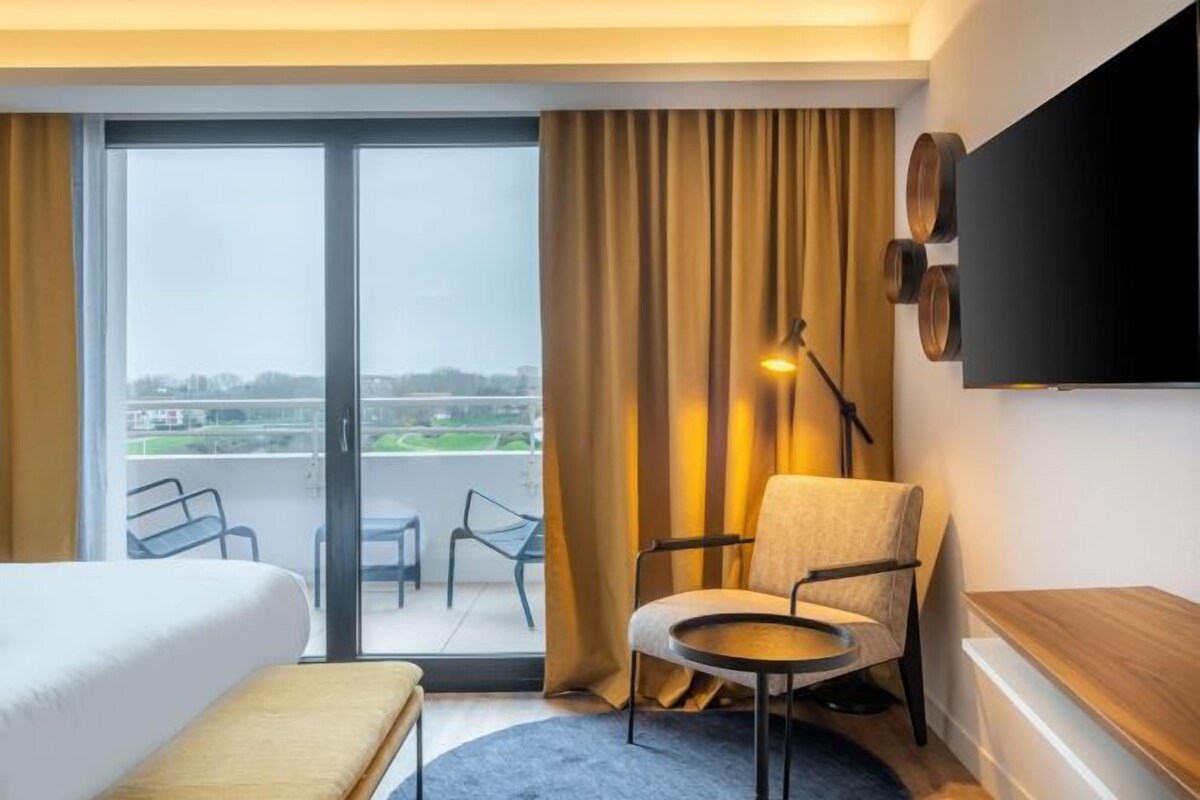 A hotel room with a bed, a chair, a small table, a TV, and a balcony with a view of the city.