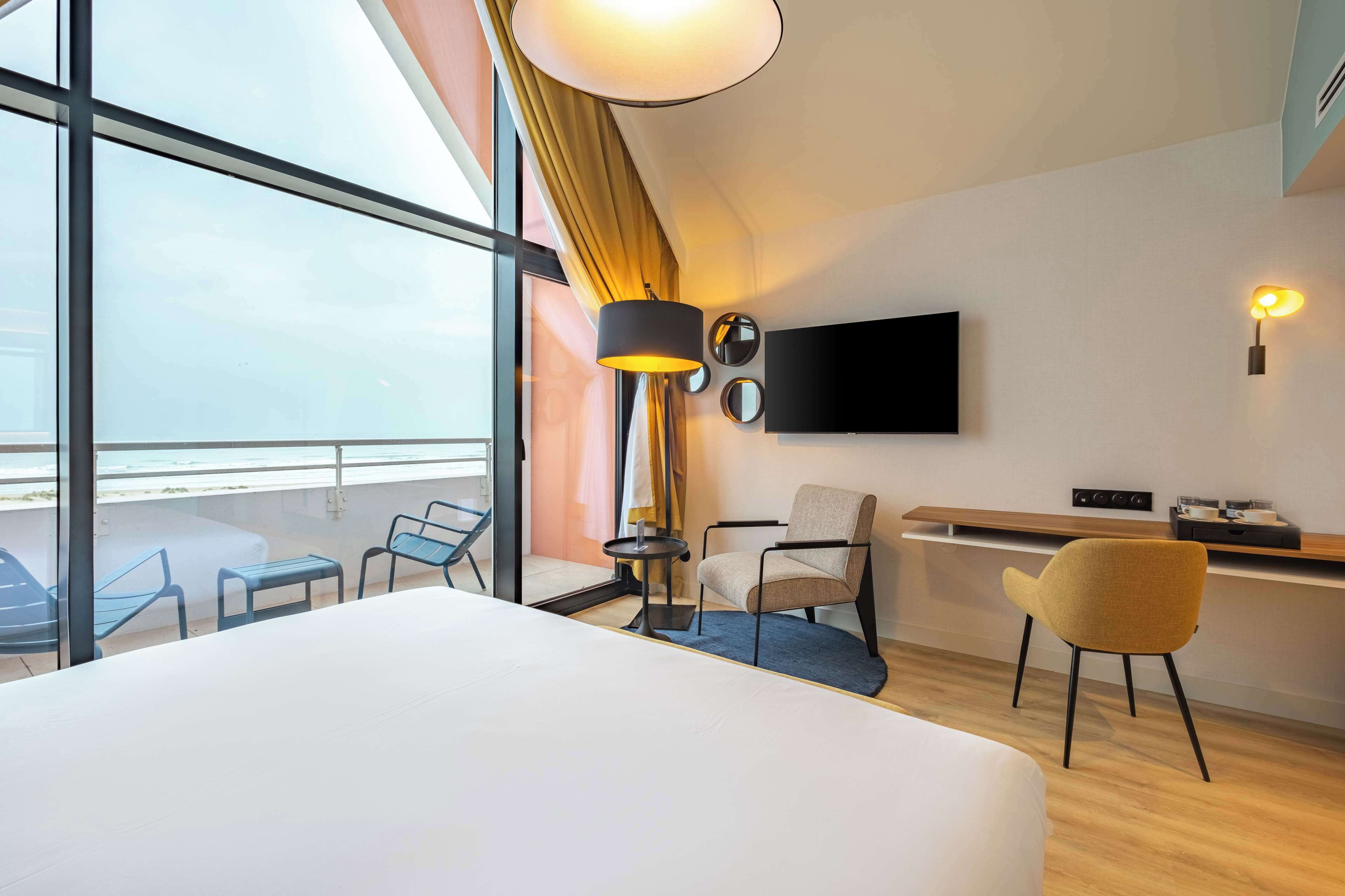 A hotel room with a large bed, a desk with a chair, a TV mounted on the wall, and a view of the sea through large windows.