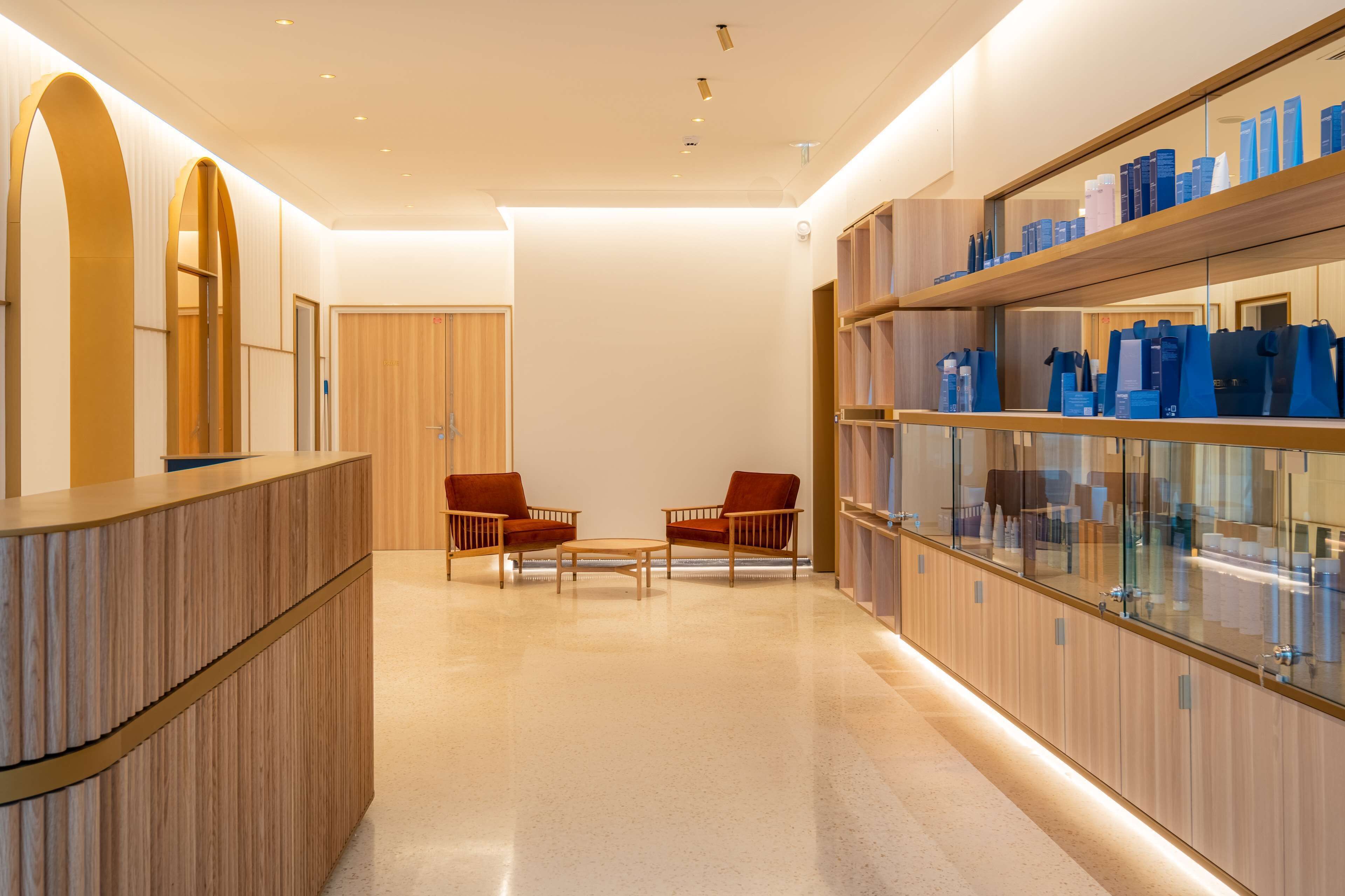 A modern hotel lobby with a wooden reception desk, two armchairs, a glass display case with toiletries, and a wooden door in the background.