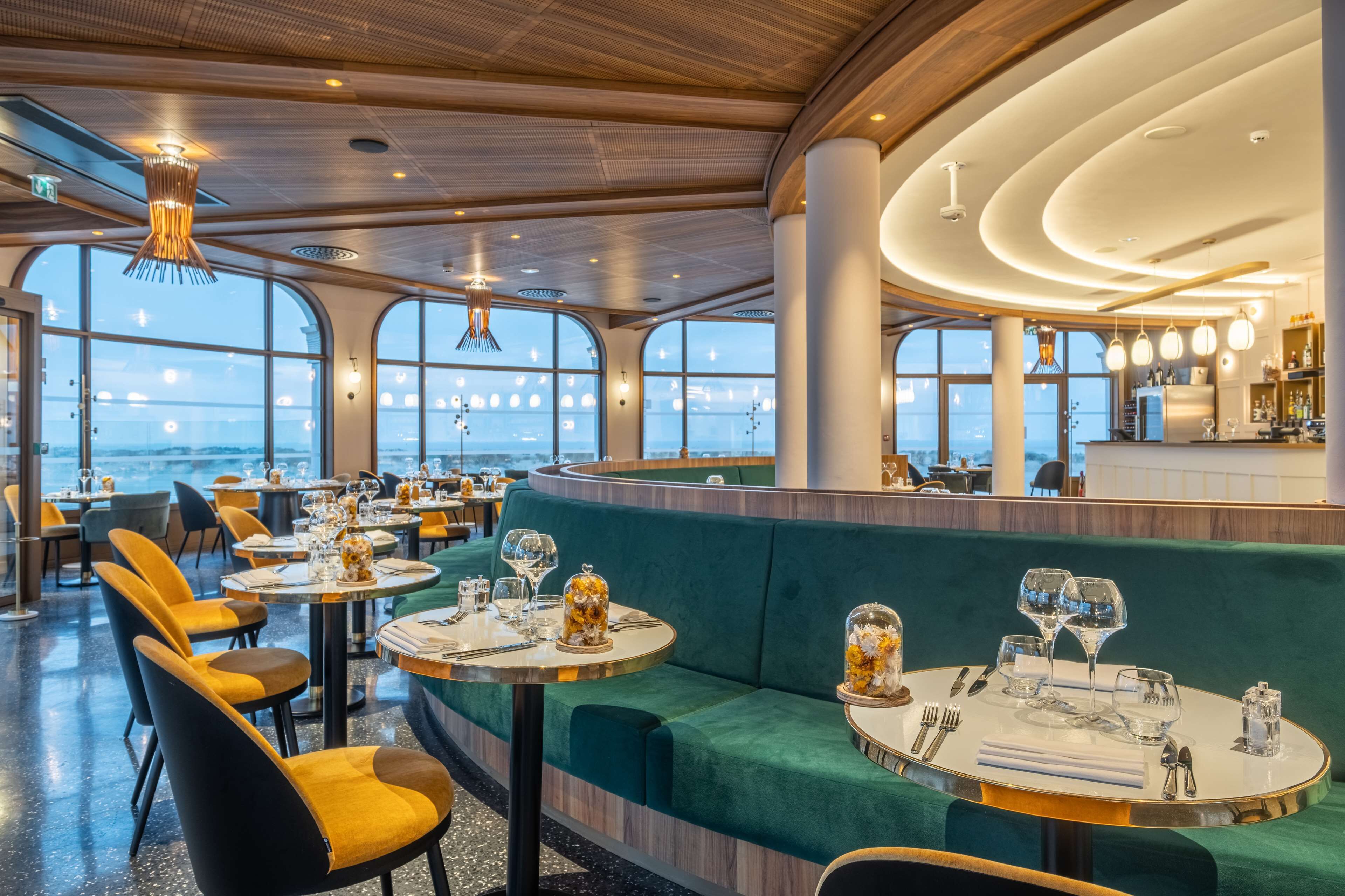 Een restaurant met ronde tafels, groen bekleden zitplaatsen en grote ramen met uitzicht op de oceaan.