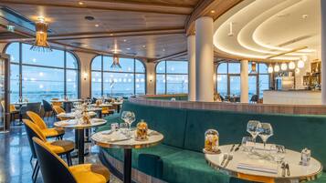 Een restaurant met ronde tafels, groen bekleden zitplaatsen en grote ramen met uitzicht op de oceaan.