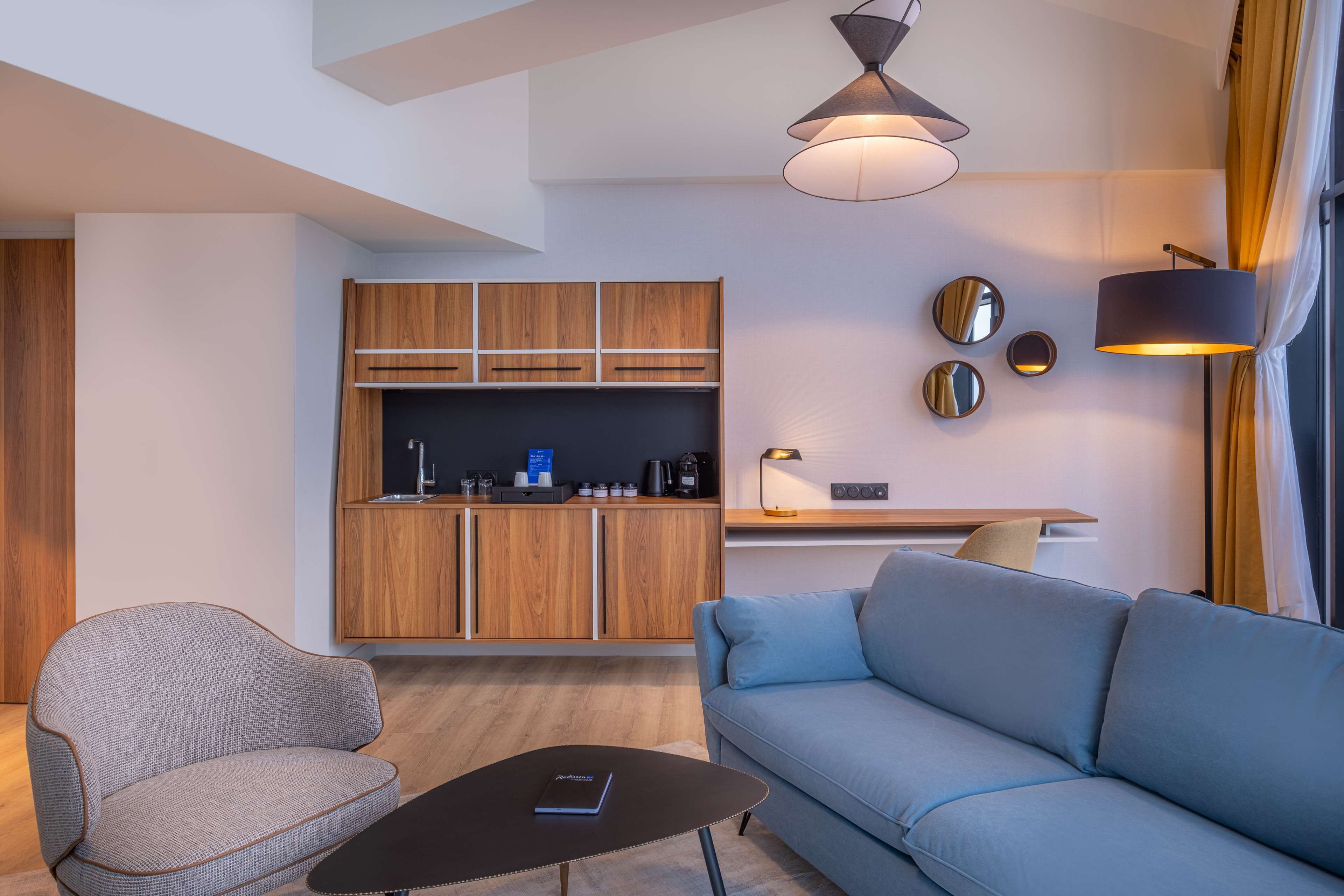 A modern living room with a sofa and armchair, a coffee table, and a kitchen area with wooden cabinets and a countertop.
