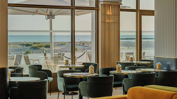 A restaurant with large windows offering a view of the beach and ocean. Tables are arranged with chairs, and there are umbrellas providing shade. - Radisson Blu Grand Hotel & Spa, Malo-Les-Bains (Dunkirk)