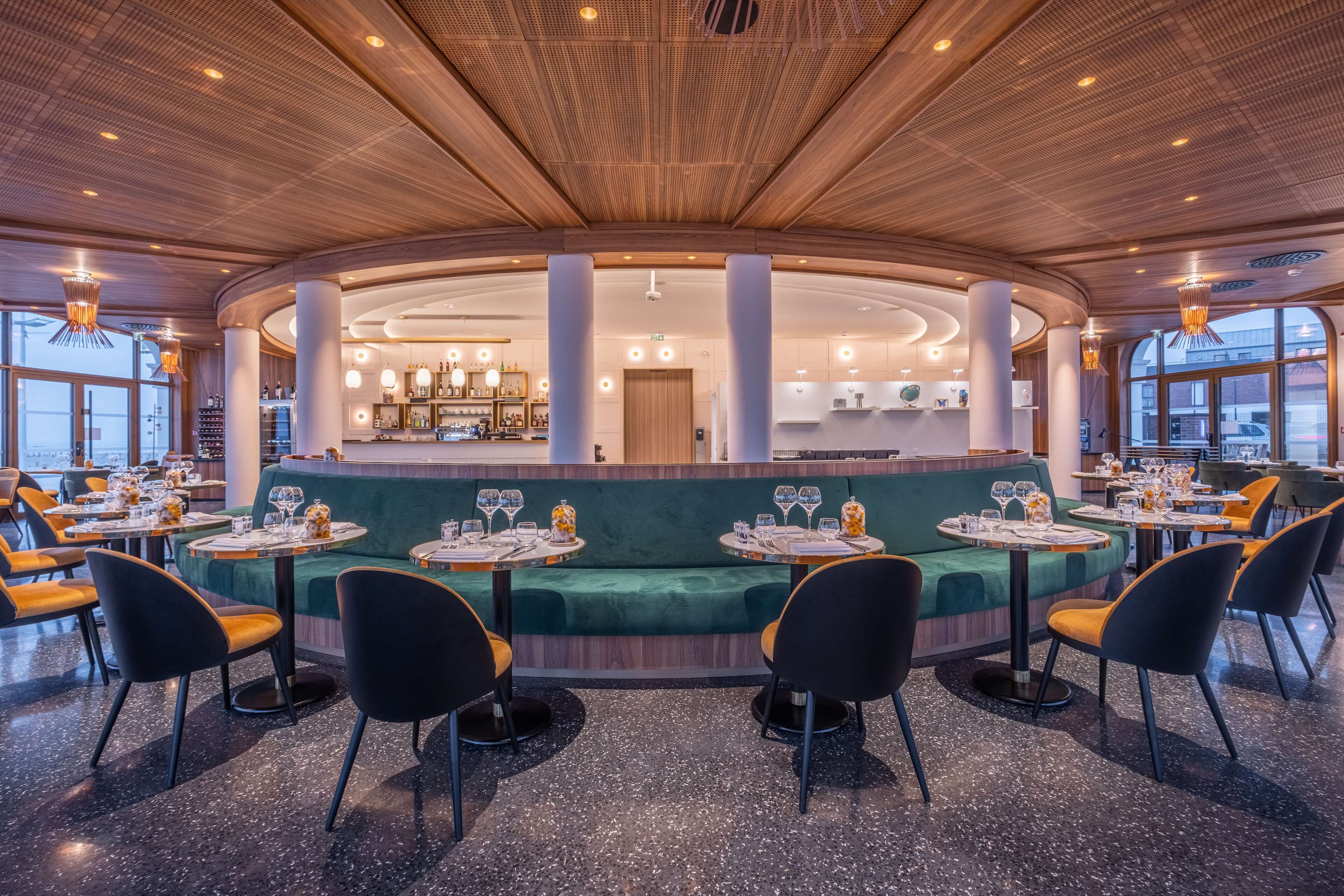 A modern restaurant with round tables, green cushioned seating, and a wooden ceiling.