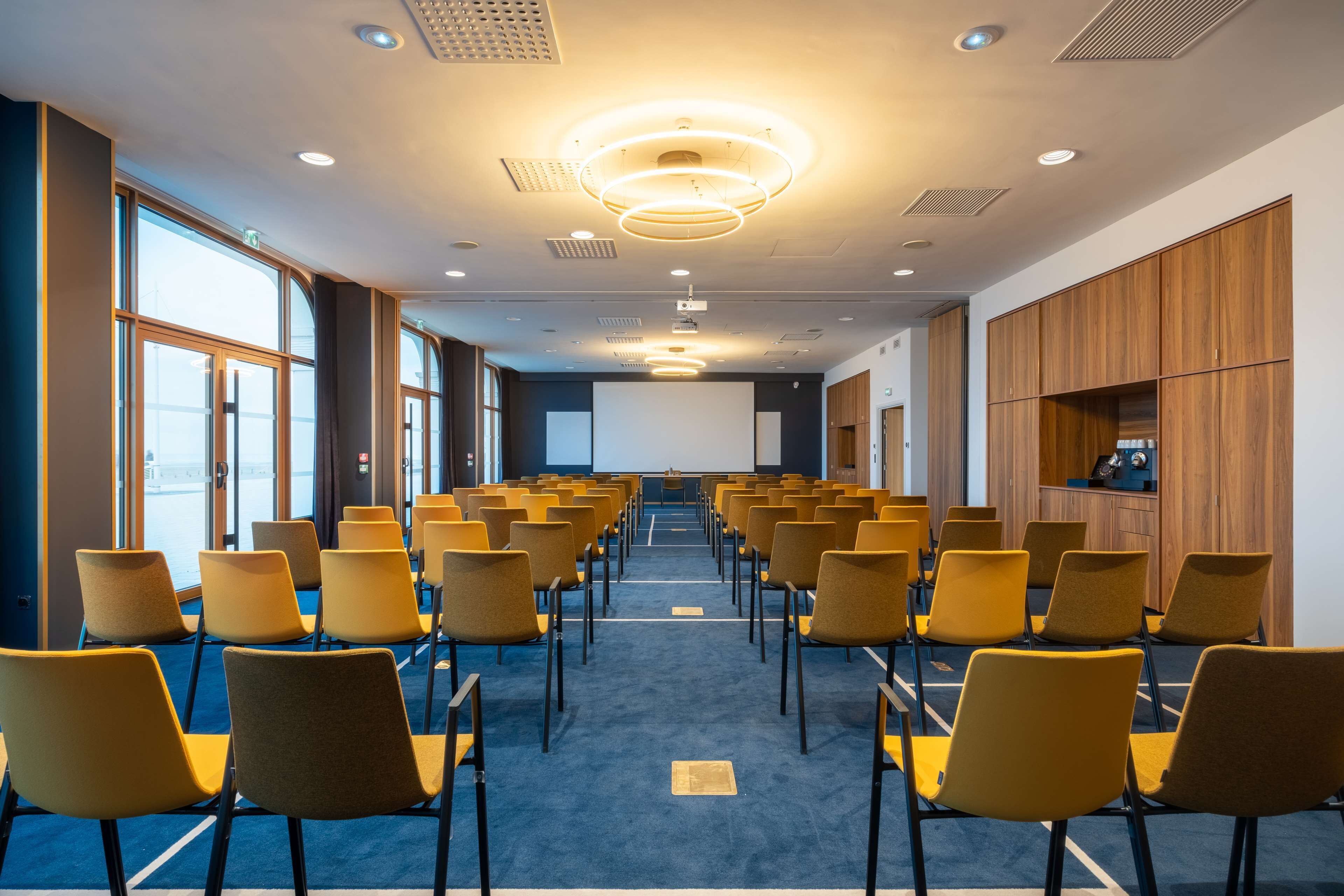 Un salón de conferencias con hileras de sillas amarillas, una pantalla grande al frente y paneles de madera en las paredes.