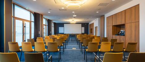A conference room with rows of yellow chairs, a large screen at the front, and wooden paneling on the walls.