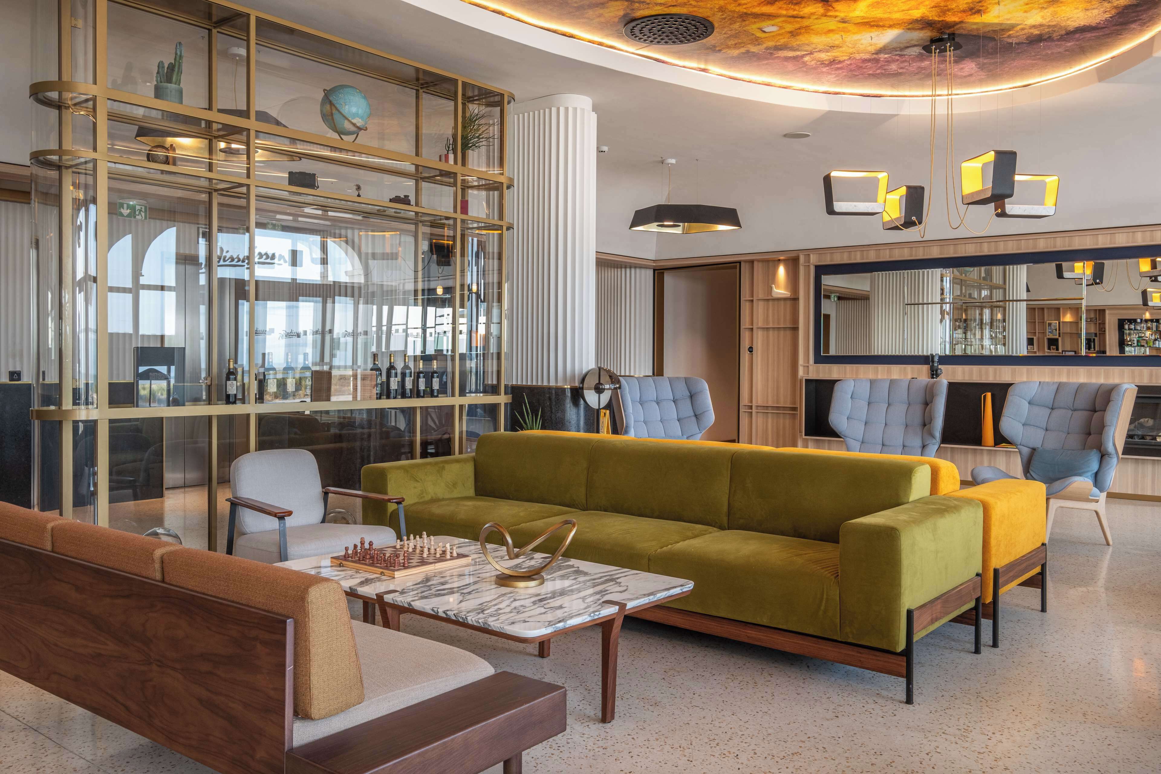 A modern living room with a green sofa, a wooden bench, and a marble coffee table. There is a glass display cabinet with various items and a large mirror on the wall.