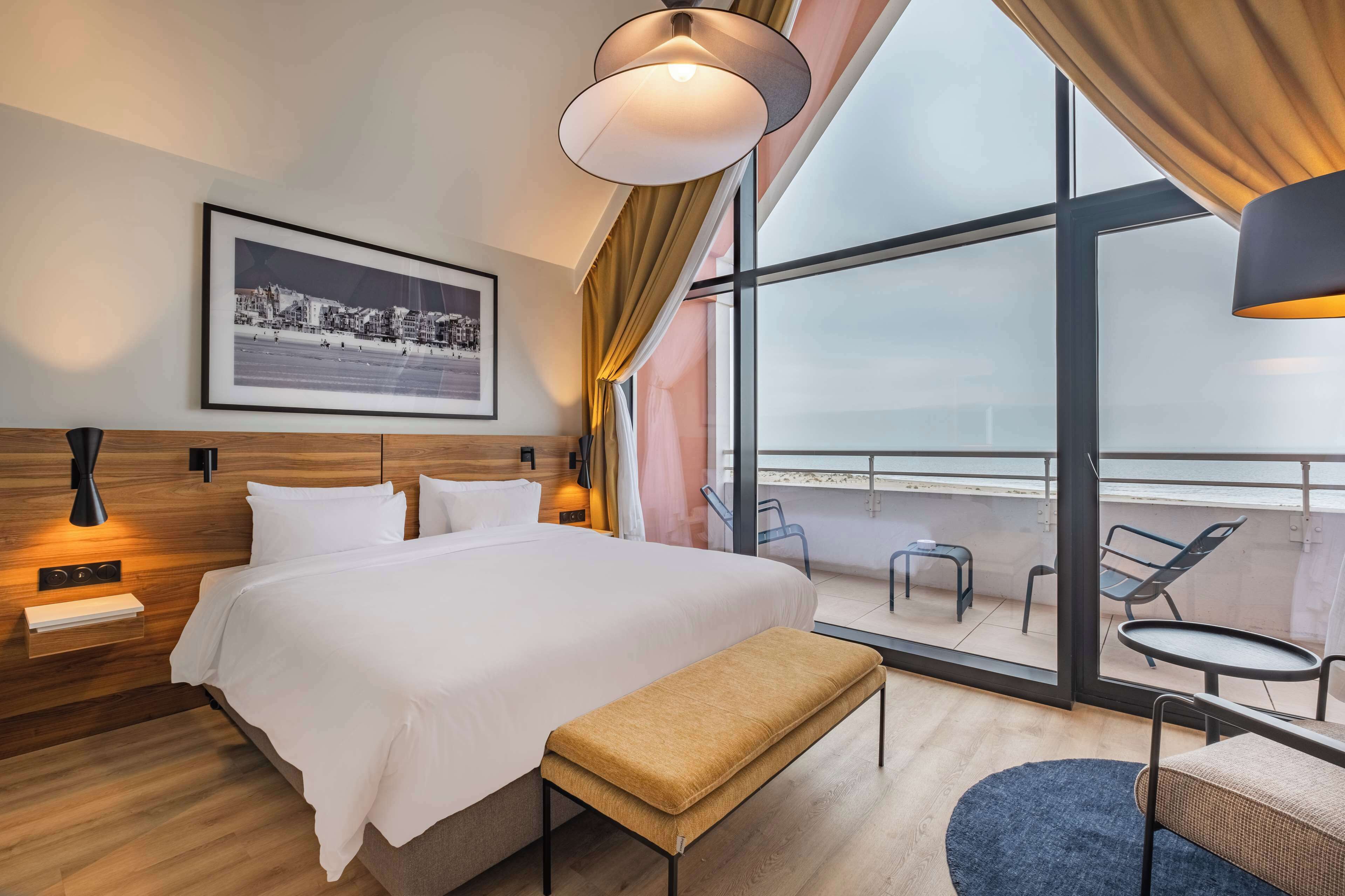 A modern hotel room with a large bed, a bench, a small table, and a balcony with chairs.