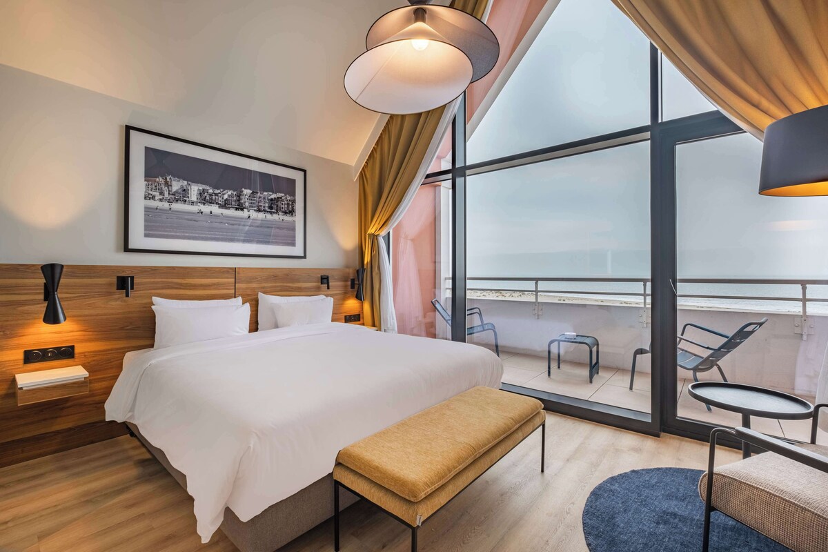 Een moderne hotelkamer met een groot bed, een bankje, een kleine tafel en een balkon met stoelen.