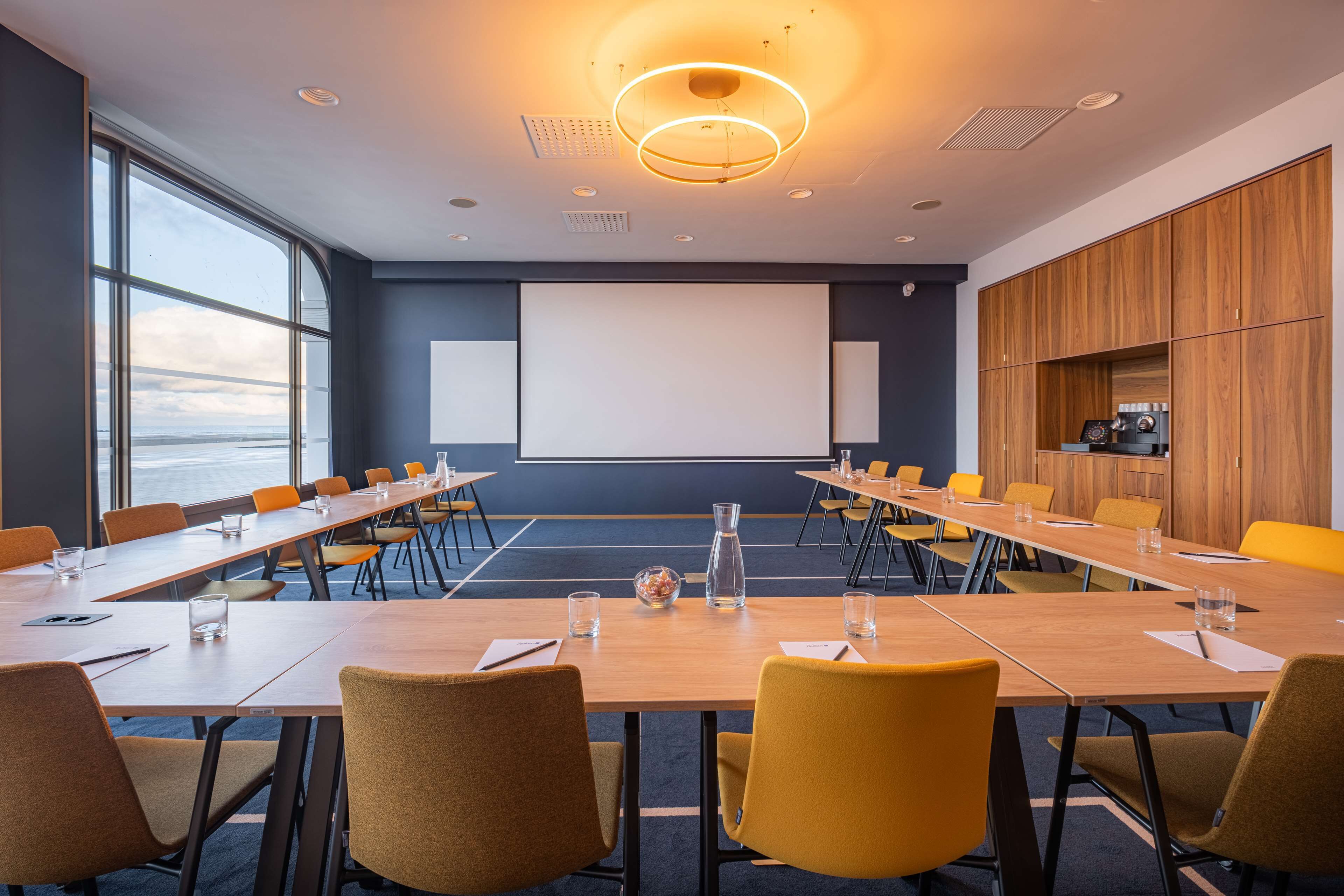 A conference room with a large screen, wooden tables, and yellow chairs.