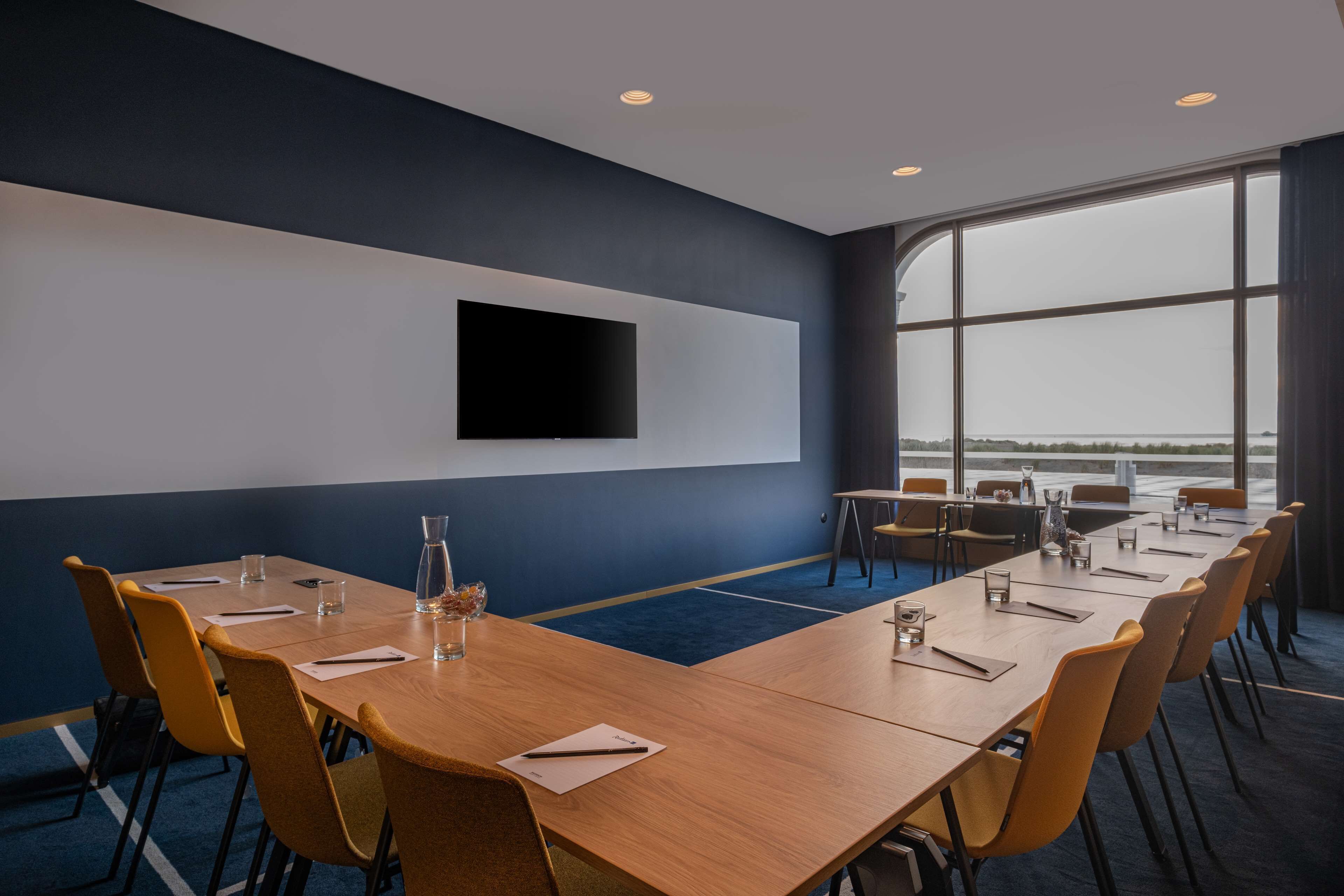 A conference room with a long table, yellow chairs, a flat-screen TV mounted on the wall, and large windows offering a view of the outdoors.