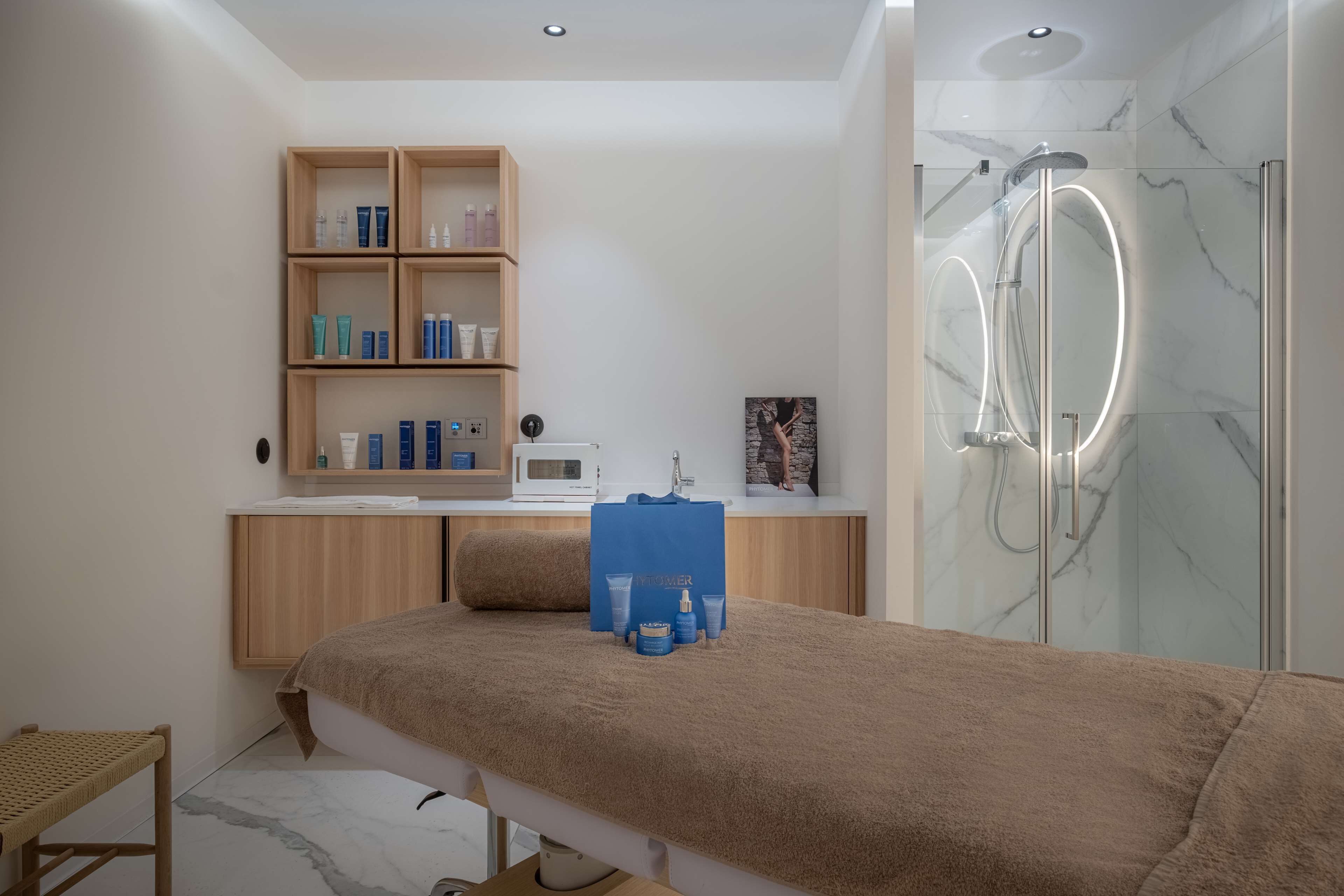 A modern spa room with a massage table, a wooden cabinet with skincare products, a shower with a glass enclosure, and a small seating area.