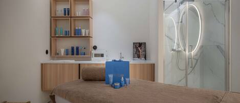 A modern spa room with a massage table, a wooden cabinet with skincare products, a shower with a glass enclosure, and a small seating area.