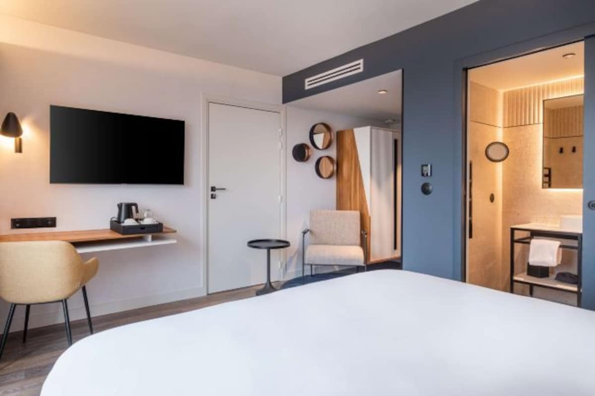 Ein modernes Hotelzimmer mit einem großen Bett, einem Schreibtisch mit Flachbildfernseher, einem Sessel, einem kleinen Tisch und einem durchgehenden Türrahmen einsehbarer Badezimmers.