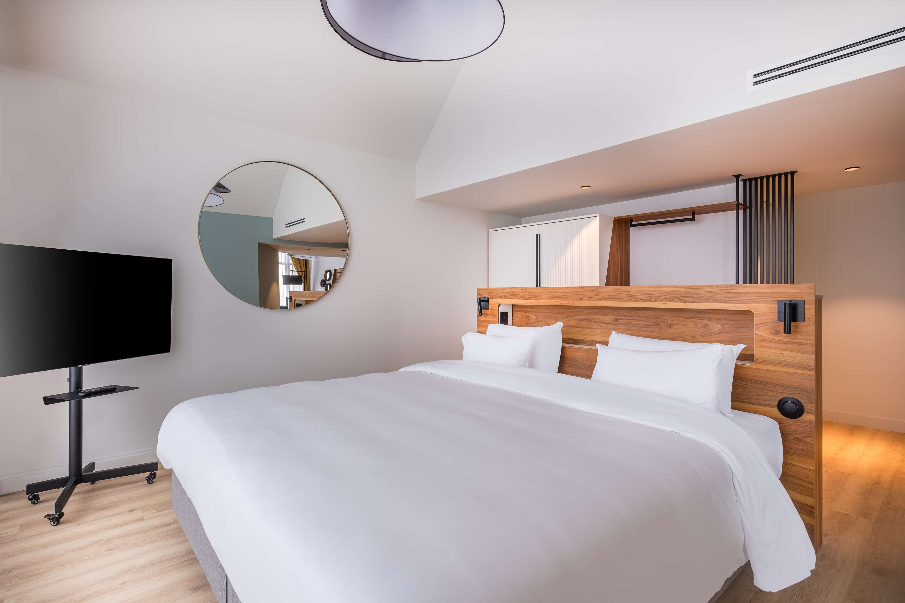 Quarto de hotel moderno com uma cama grande, televisão de tela plana, espelho redondo e cabeceira de madeira.