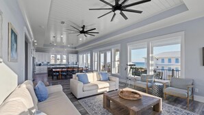 Amplia sala de estar con sofá, mesa de centro y sillones, una cocina al fondo y ventanas grandes con vista al océano.