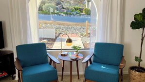 Une pièce avec deux fauteuils bleus, une petite table ronde avec une lampe, une plante en pot et une vue sur un jardin par une grande fenêtre.