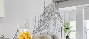 Una sala de estar moderna con un gran cuadro arquitectónico blanco y negro, un sofá cómodo, una mesa de comedor con una bolsa de palomitas de maíz amarillas y una planta en maceta.