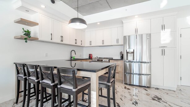 A modern kitchen with white cabinets, a central island with bar stools, stainless steel appliances, and a pendant light. - The Caïman Complex, 707 + Parking (Québec City)