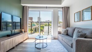 Ein modernes Wohnzimmer mit Flachbildfernseher, einem grauen Sofa, einem kleinen runden Tisch und einem großen Fenster mit Blick auf die Stadt.