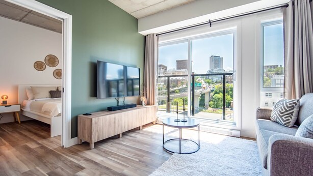 Ein modernes Wohnzimmer mit einem Bett, einem Fernseher, einem runden Tisch und großen Fenstern mit Blick auf die Stadt.