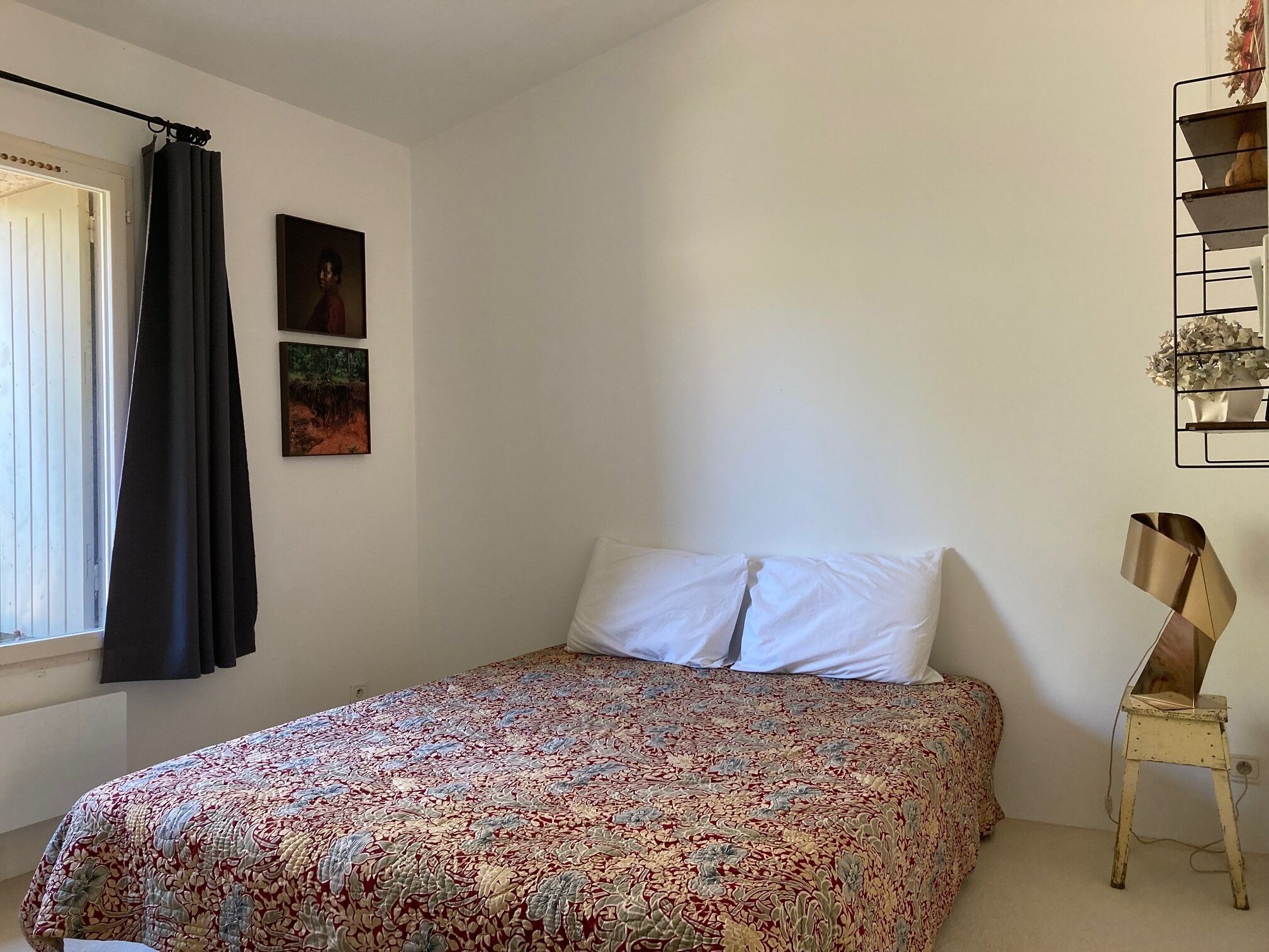 Un dormitorio con cama, ventana con cortinas, estante en la pared con flores y un cuadro.
