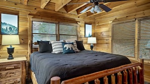 Une chambre à coucher dans une cabane en bois, avec un lit, deux fenêtres équipées de stores, un ventilateur de plafond et une table de chevet avec une lampe.