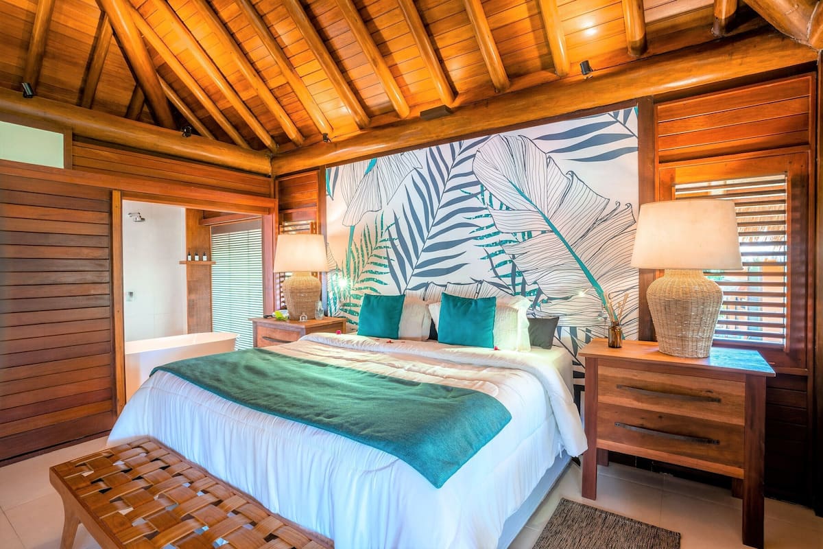 Un plafond en bois, un lit recouvert d’une couverture verte, deux tables de chevet avec des lampes et un mur orné d’un motif de feuilles.