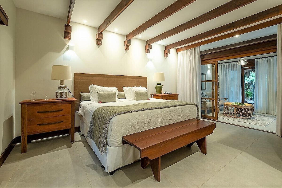 Une chambre avec un grand lit, une tête de lit en bois, des tables de chevet avec des lampes, un banc et une vue sur l’extérieur grâce à une porte vitrée.