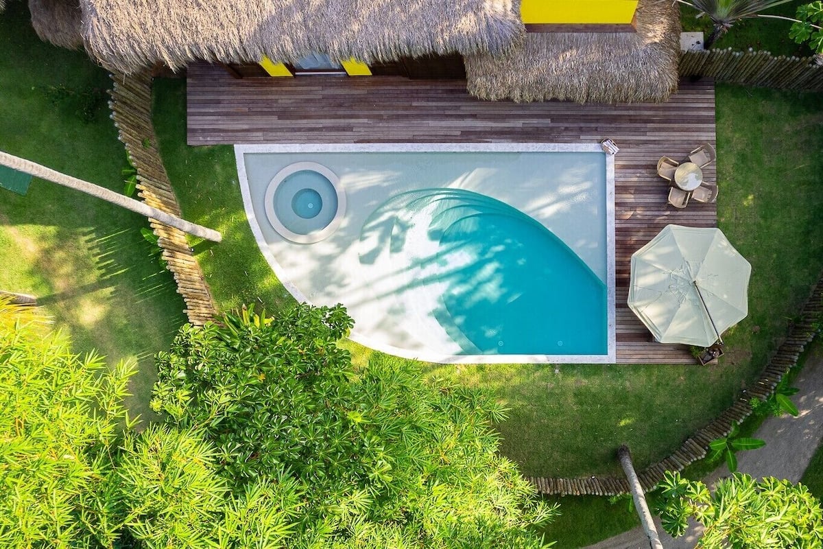Vue aérienne d’une piscine dotée d’une terrasse en bois, entourée d’une végétation luxuriante et bordée d’une structure au toit de chaume.
