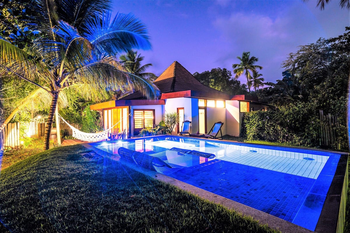 Une maison avec une piscine, des palmiers et un hamac.