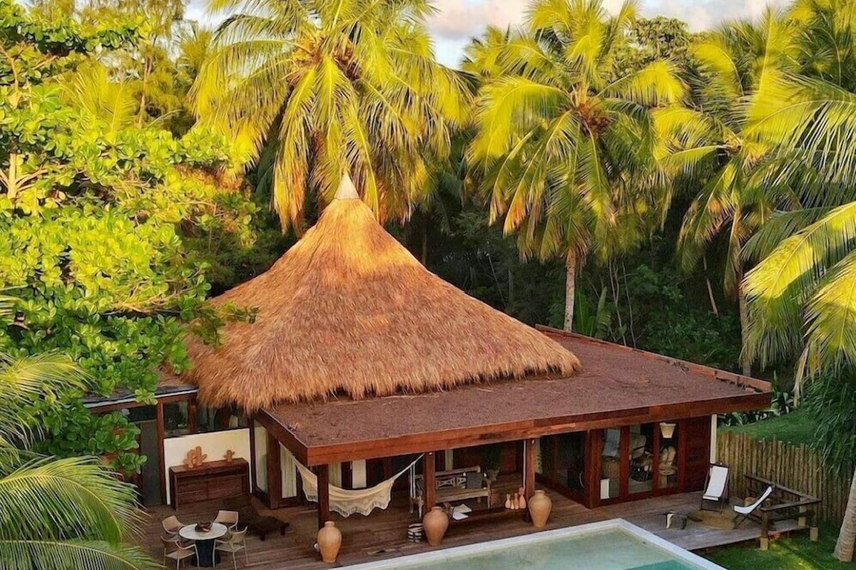 Une maison à toit de chaume, dotée d’une piscine et d’un espace extérieur aménagé, le tout entouré de palmiers.