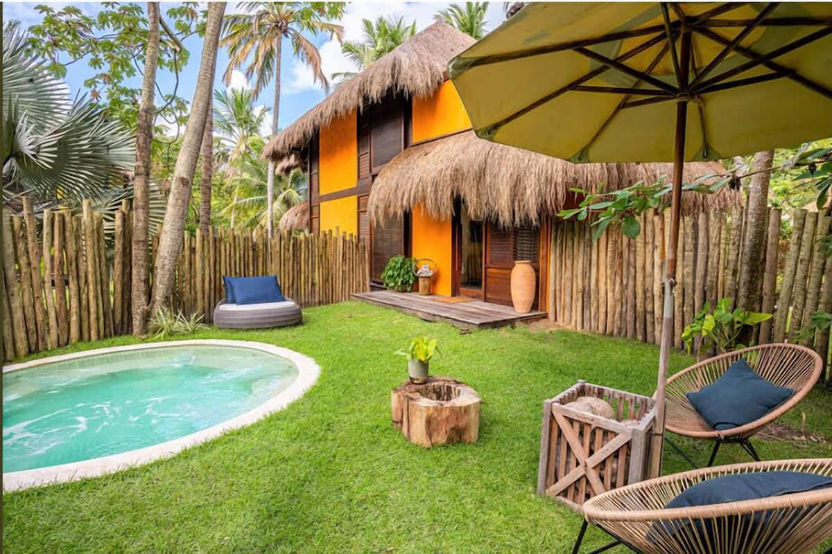 Une maison à toit de chaume avec une piscine, des meubles en osier et une clôture en bois.
