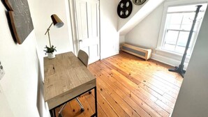 Una habitación con techo inclinado, suelo de madera, una mesita con lámpara, una puerta y una ventana con persianas.