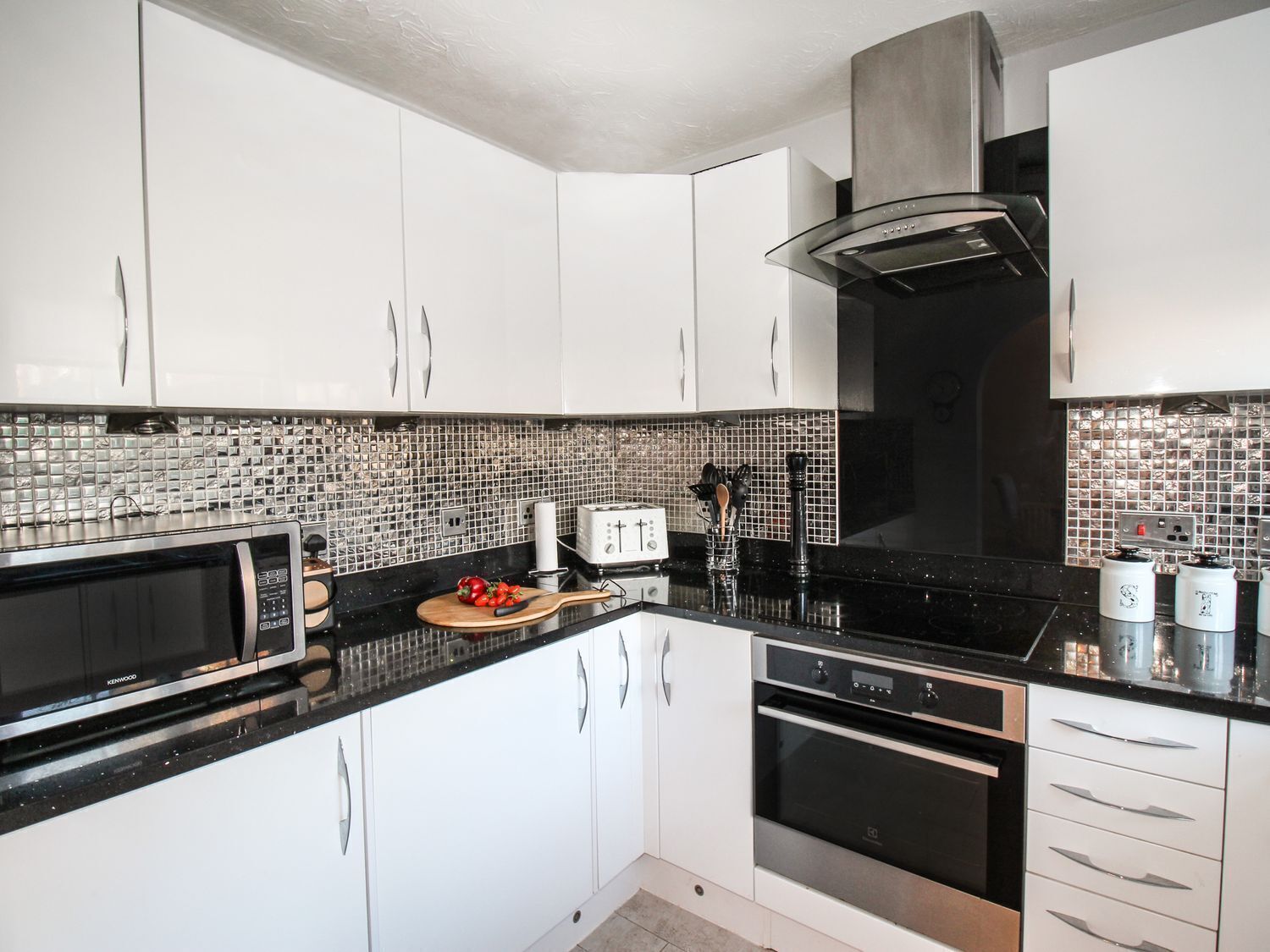 A modern kitchen with white cabinets, a black countertop, a Kenwood microwave, a toaster, and a stainless steel range hood.