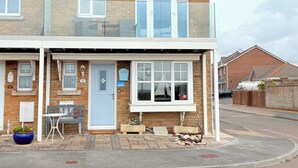 Une maison à deux étages avec un porche à l’avant, une porte bleue et un petit espace extérieur aménagé avec une table et des chaises.