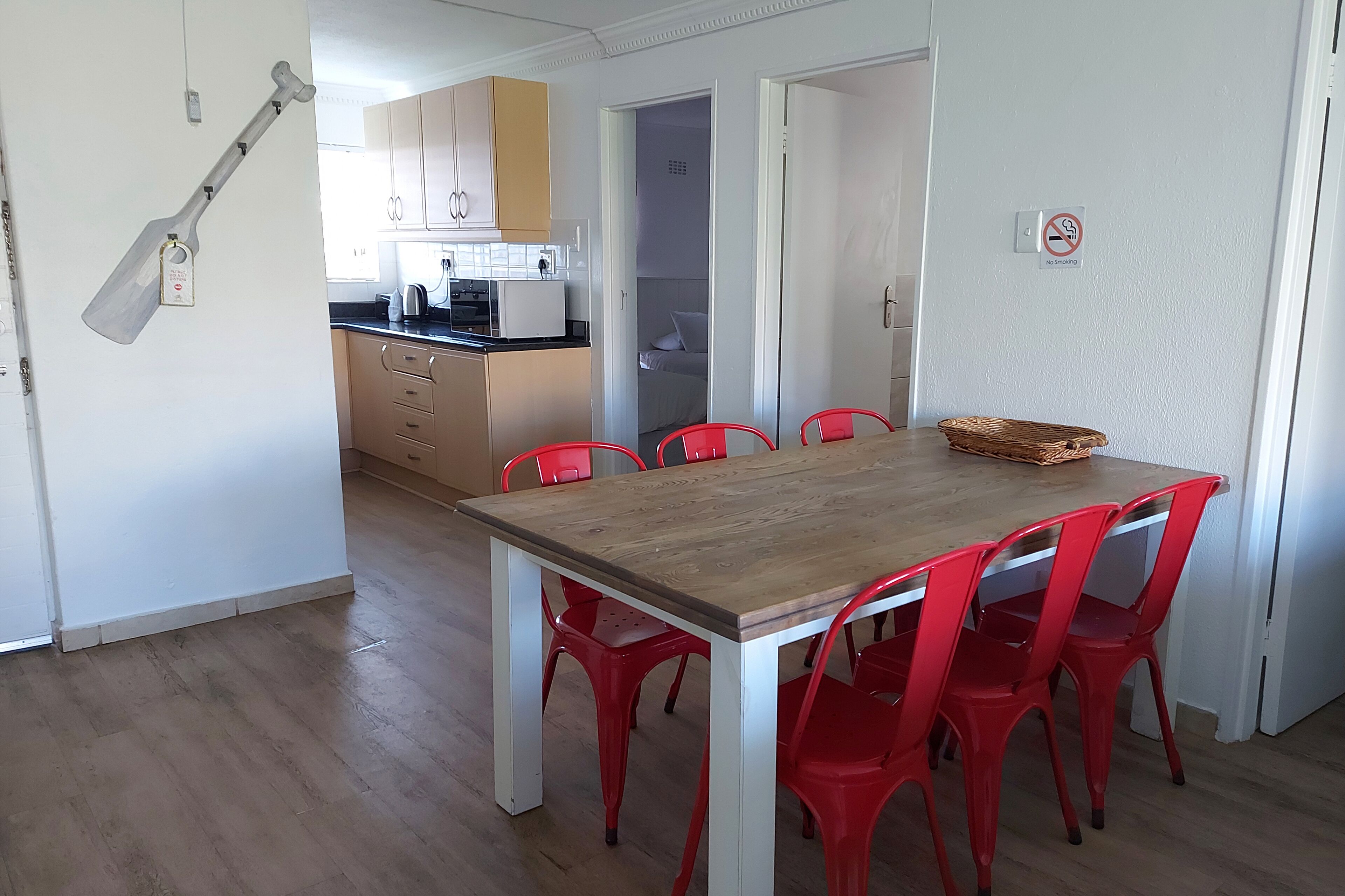Un comedor con mesa de madera y sillas rojas, una cocina al fondo y un cartel de prohibido fumar en la pared.