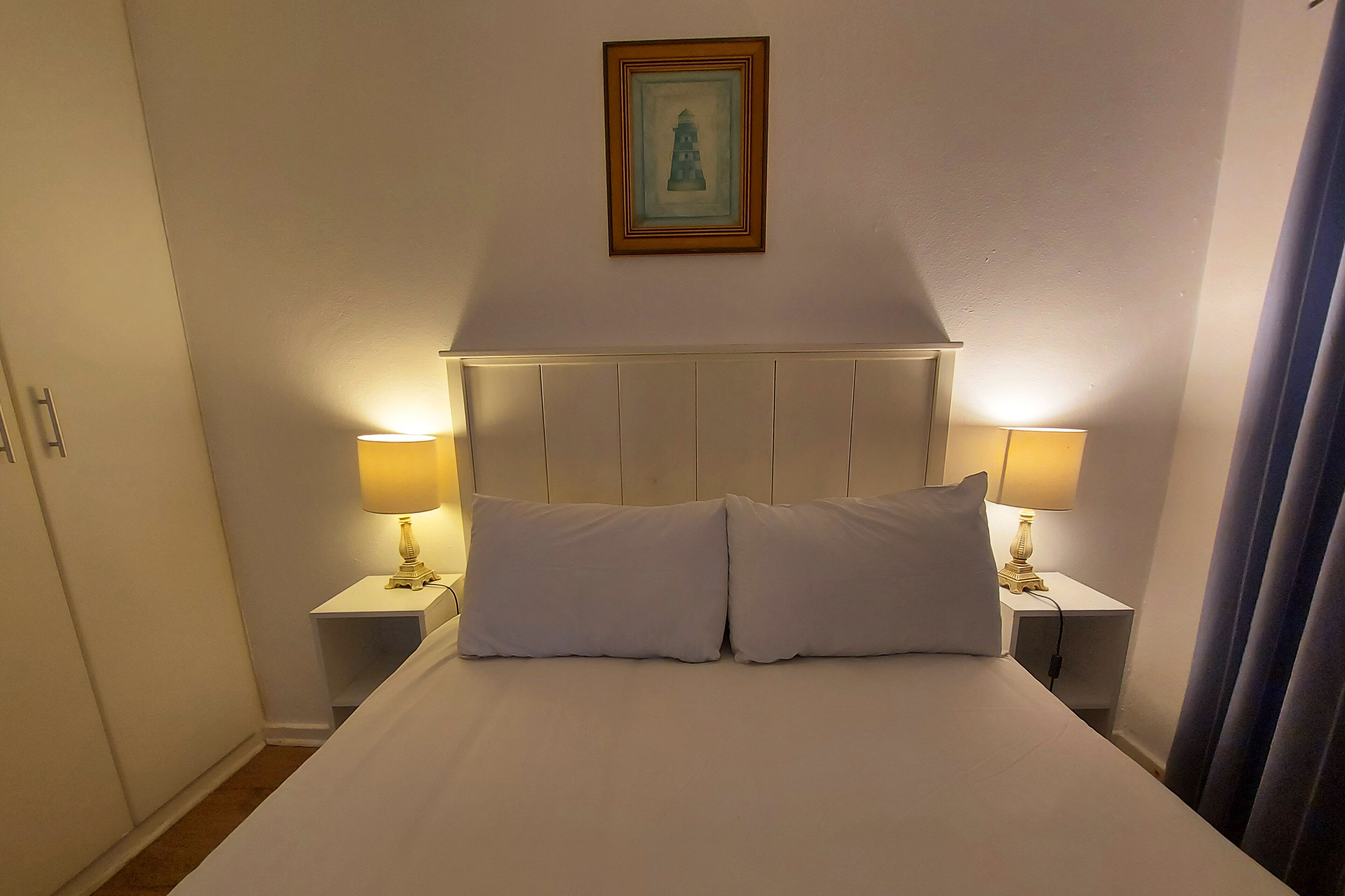 Una cama bien hecha con sábanas blancas y dos almohadas, flanqueada por mesitas de noche con lámparas. Un cuadro enmarco cuelga sobre la cama.