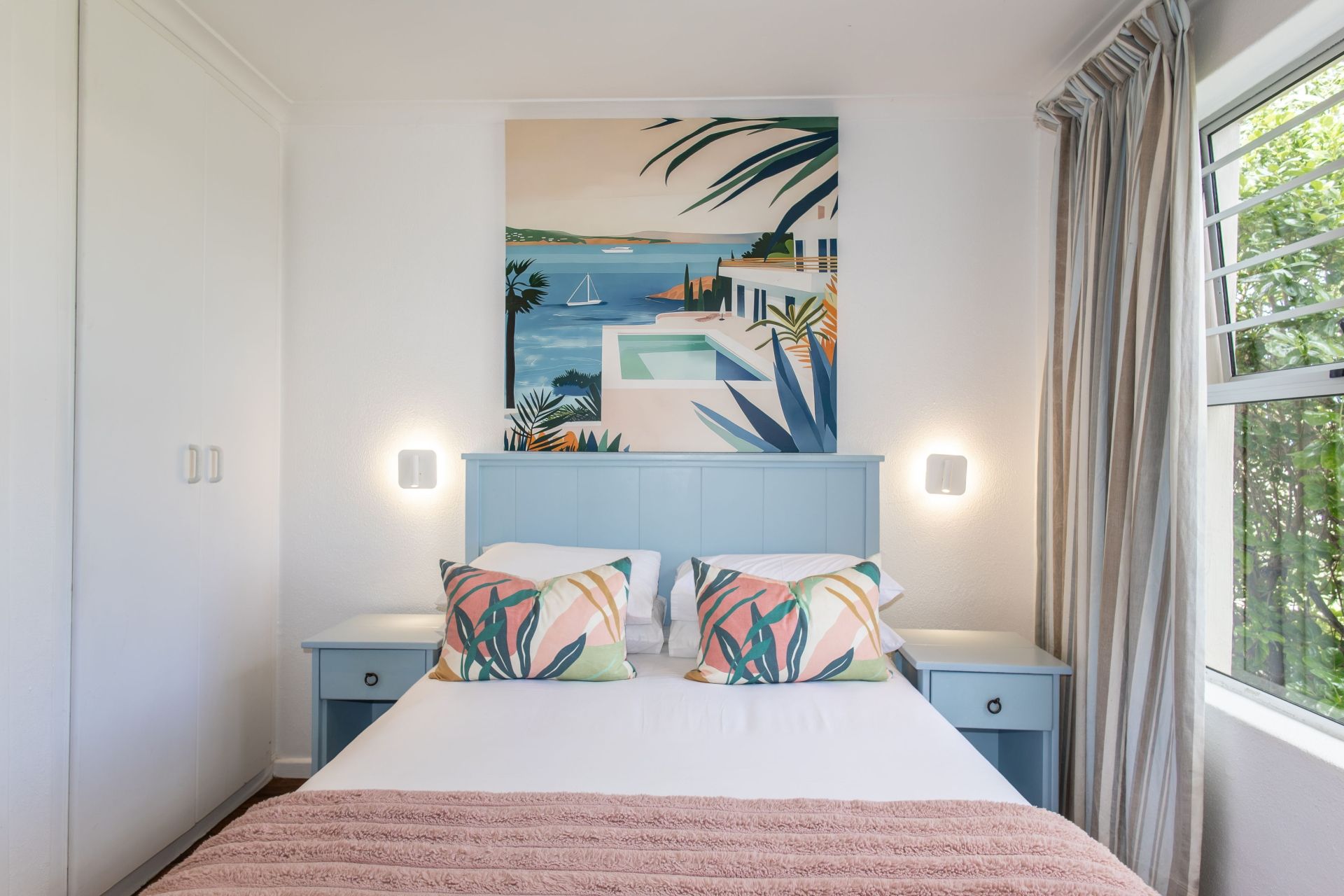 A bedroom with a bed, two bedside tables, a wall-mounted painting, and a window with curtains.