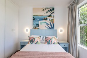 A bedroom with a bed, two bedside tables, a wall-mounted painting, and a window with curtains. - First Group Hermanus Beach Club (Hermanus)
