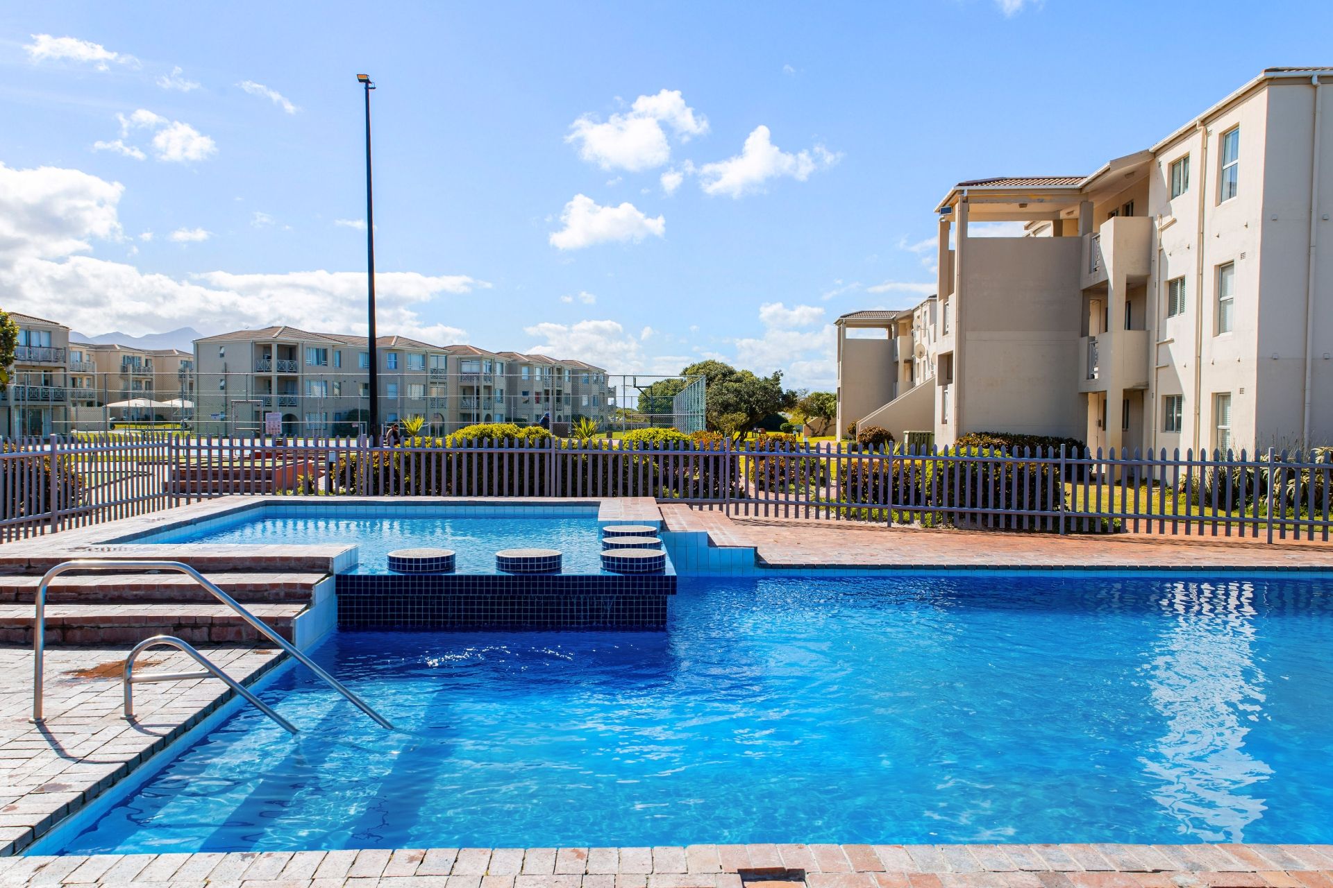 Una piscina con una escalera metálica y escalones, rodeada por una valla, con edificios de apartamentos al fondo.