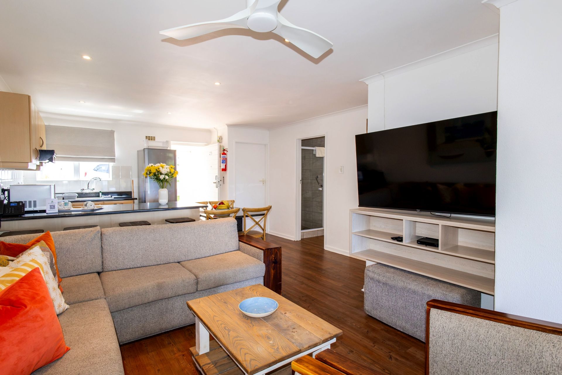 A modern living room with a flat-screen TV, grey sofas, a wooden coffee table, and a ceiling fan.