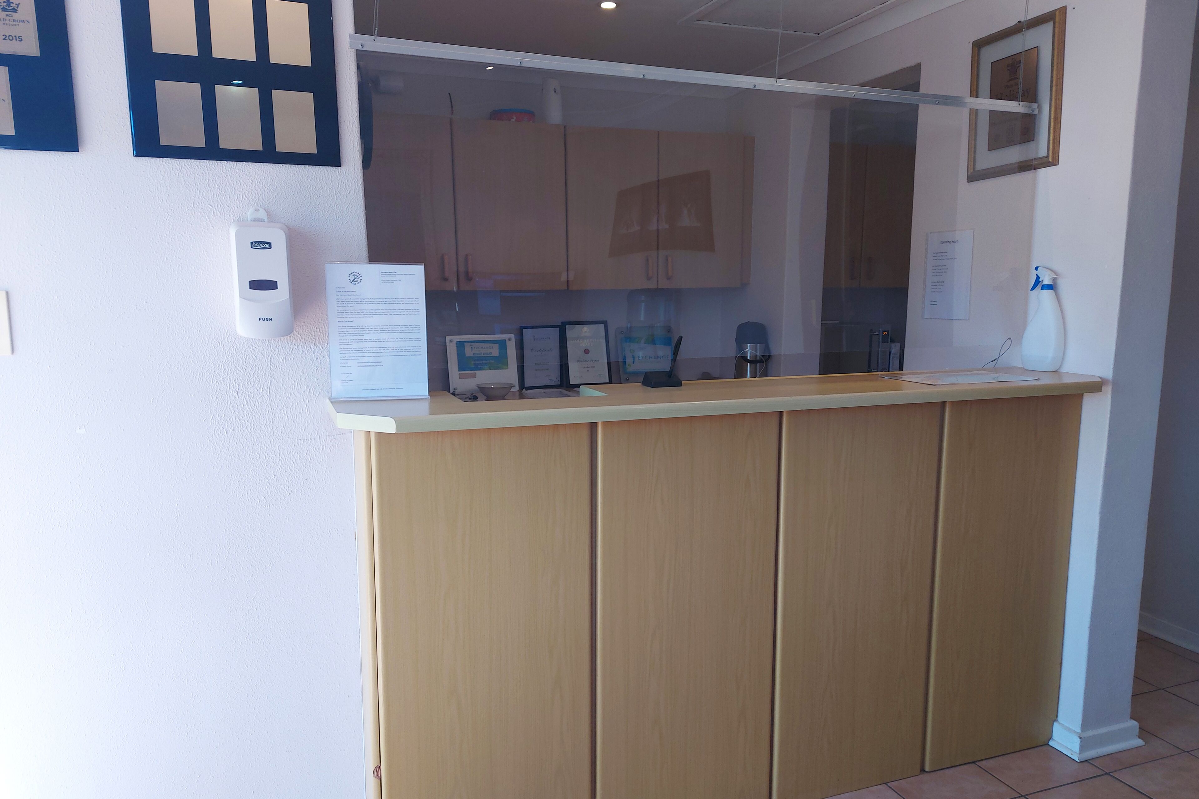 A reception desk with a glass partition, wooden cabinets, a white wall-mounted device, and a framed certificate on the wall.