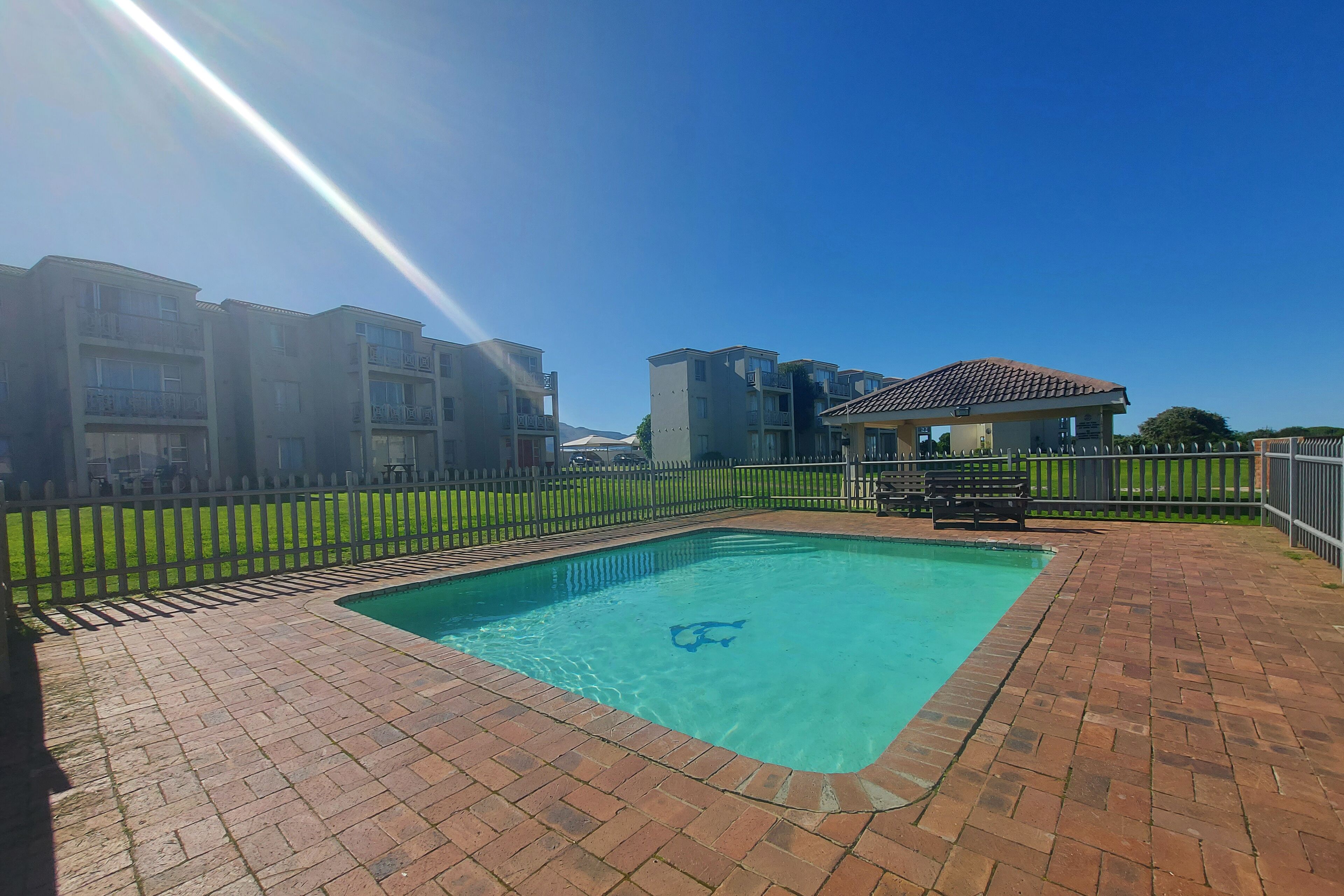 Une piscine rectangulaire avec une terrasse carrelée, entourée d’une clôture blanche en bois, et située à côté d’un immeuble résidentiel à plusieurs étages.