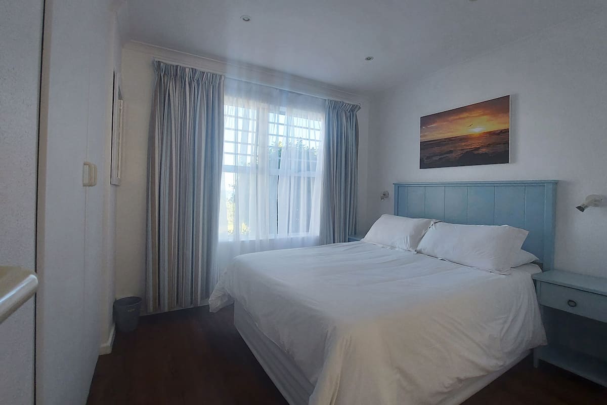 Un dormitorio con cama, mesita de noche, un cuadro de una puesta de sol y una ventana con cortinas.