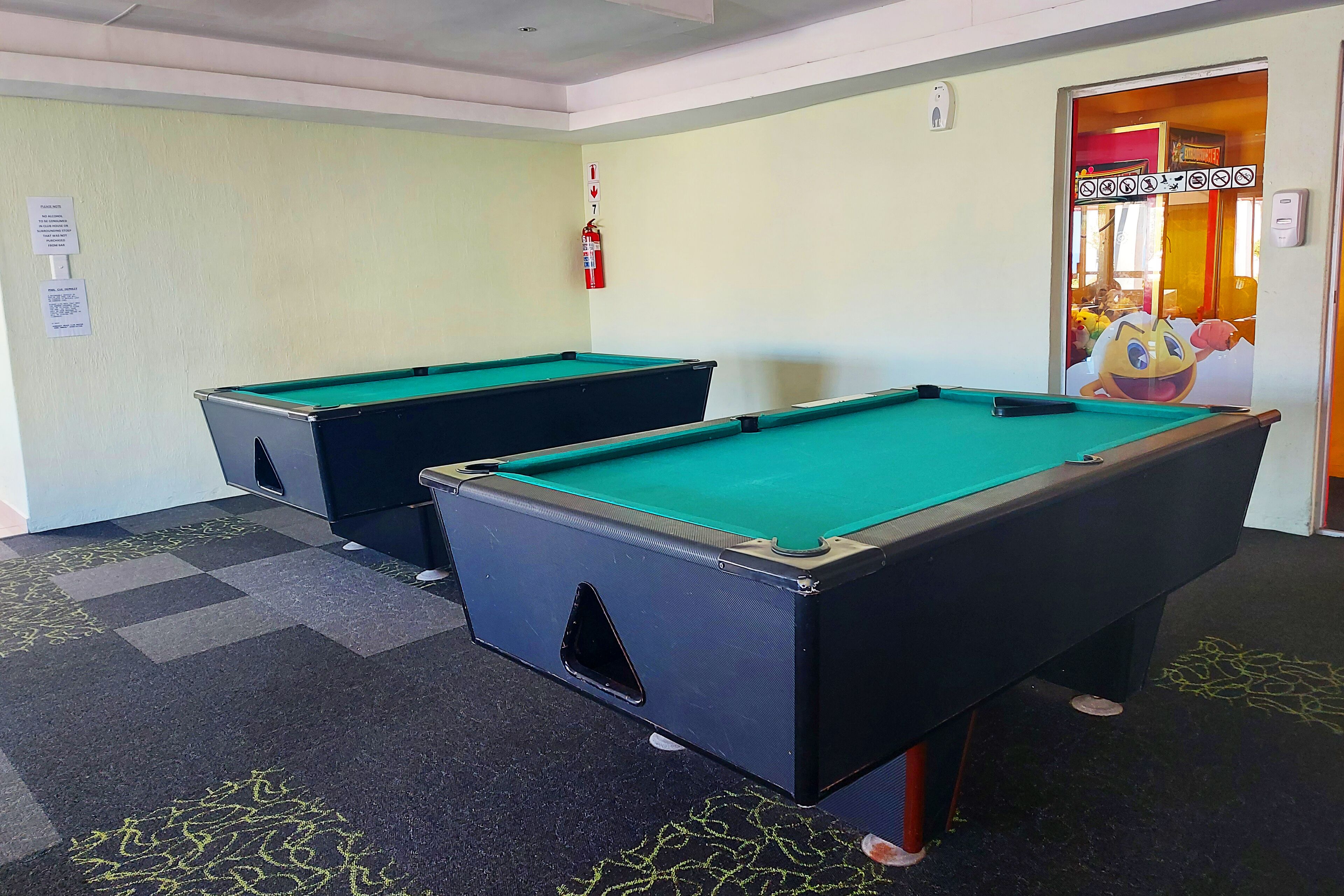 Two pool tables in a room with a fire extinguisher on the wall and a door leading to another area.